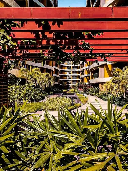The landscaping at Palm Bellagio is designed to complement the architecture. This view through a red pergola frames the building's unique facade, showing how we integrate green spaces and structural elements to create a cohesive environment.