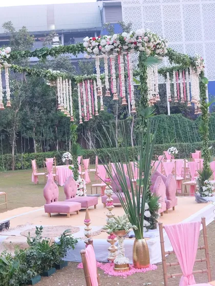A beautiful pink and green mandap designed for an outdoor wedding ceremony. The setup includes lush greenery, hanging floral arrangements of tuberose and roses, and elegant pink seating for the family.
