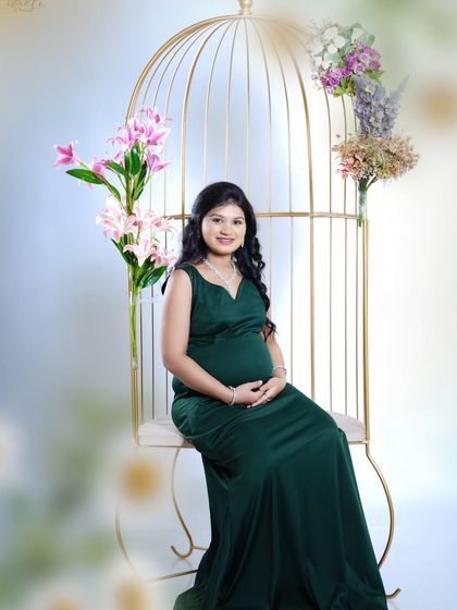 A serene solo portrait of the mother-to-be seated in our golden birdcage chair. She is wearing a beautiful, dark green fitted gown that elegantly showcases her pregnancy silhouette.