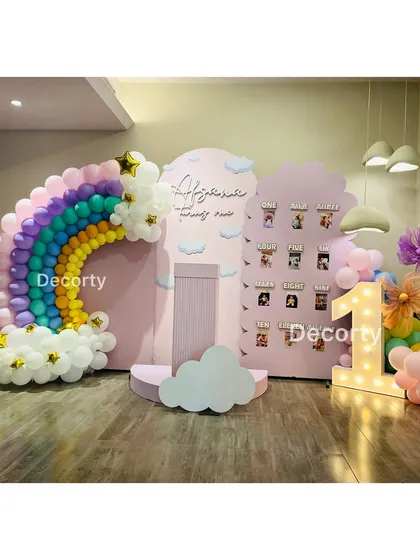 A magical rainbow and cloud-themed backdrop for a first birthday. The setup includes a balloon rainbow, a "My First Year" photo display, and a large light-up number one.