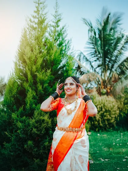 A beautiful outdoor portrait showcasing a traditional South Indian bridal look. The soft, natural sunlight brings out the vibrant orange and white of the saree and the warmth of the gold jewelry, capturing a moment of pure joy.