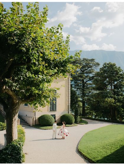 A couple stands in the manicured gardens of an Italian villa, with mountains in the background. The image is peaceful and elegant, showcasing the beauty of a Lake Como pre-wedding shoot.