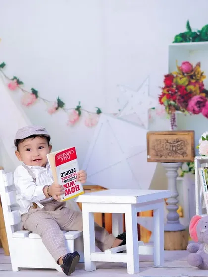 A one-year-old boy in a charming outfit with a cap, sitting at a small table and holding a book in our bright studio setup.