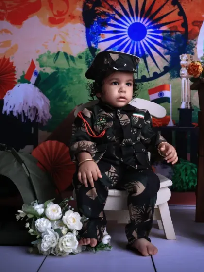 A young child dressed in a full army camouflage uniform for an Independence Day themed shoot. He sits seriously on a small chair, embodying the spirit of a future soldier, surrounded by patriotic props.