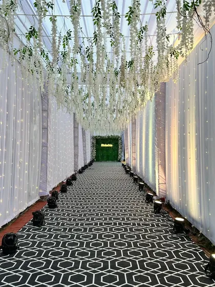 A long view of the Sangeet passage, with its ceiling of hanging wisteria and a pathway lined with lights, leading to a green photo wall at the end.