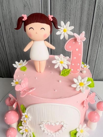 A sweet and simple first birthday cake in baby pink. This design is adorned with tiny butterflies, flowers, mushrooms, and a cute, hand-sculpted doll topper, creating a magical and charming centerpiece for a little one's special day.