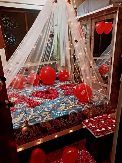 A cozy first-night room decoration with a canopy of fairy lights over the bed. The bed is covered in rose petals and red balloons to complete the romantic theme.
