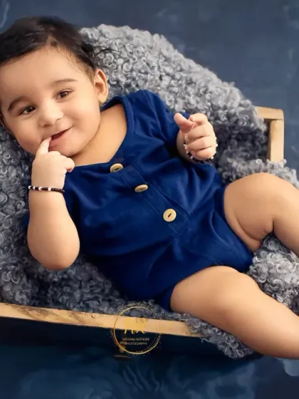 A baby boy in a blue romper lies in a wooden box, playfully putting his finger in his mouth.