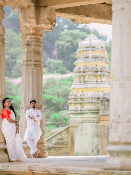 A wide shot of a couple posing amidst the stone pillars of a historic temple, with the gopuram visible in the background. This composition emphasizes the grandeur of the location.