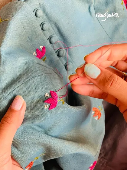 A close-up of the hand-embroidery process for our Cheenth collection. The artisan uses a satin stitch to create vibrant pink floral buds on a light blue blouse with fabric-covered buttons.