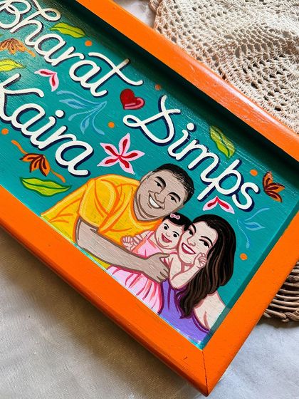 A beautiful family nameplate we painted for Dimple, featuring portraits of her, her husband Bharat, and their daughter Kaira. The bright orange frame makes the artwork pop.