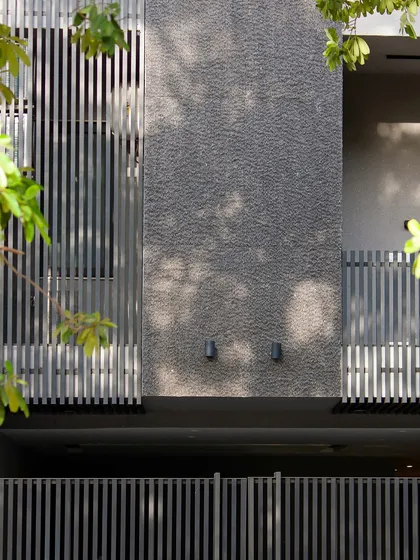 A close-up of the facade, showing the vertical lines of the metal louvers and gate against the rough texture of the wall. The play of light and shadow through the trees adds another layer of detail.