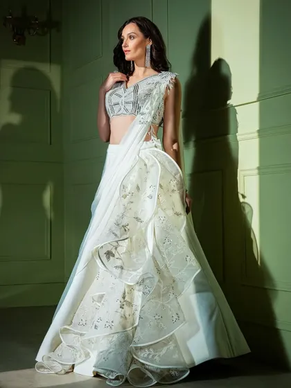 A masterpiece in white. This unique gown combines a draped skirt with a ruffled, layered organza panel, paired with a stunningly embroidered one-shoulder blouse.