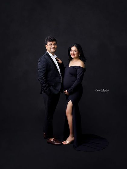 A full-length shot of a couple in elegant black attire. The off-the-shoulder gown adds a touch of modern glamour to this classic studio maternity portrait.