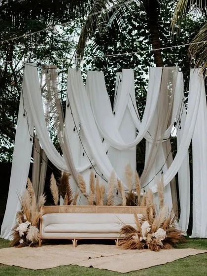 A rustic and romantic photo corner for an outdoor event. The backdrop is made of flowing white and beige drapes, with a simple wooden sofa surrounded by pampas grass and dried floral arrangements.