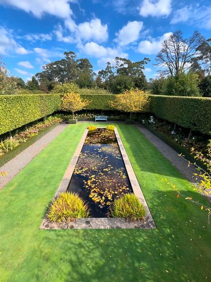 A rectangular formal pond, also known as a reflection pool, creates a stunning centerpiece. It is surrounded by a crisp lawn border and tall, dense hedges, reflecting the sky and surrounding trees.