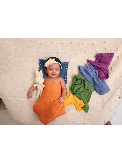A smiling newborn lies on a soft blanket, surrounded by a rainbow of wraps. This happy portrait, complete with a toy bunny, captures a moment of pure, sleepy bliss.