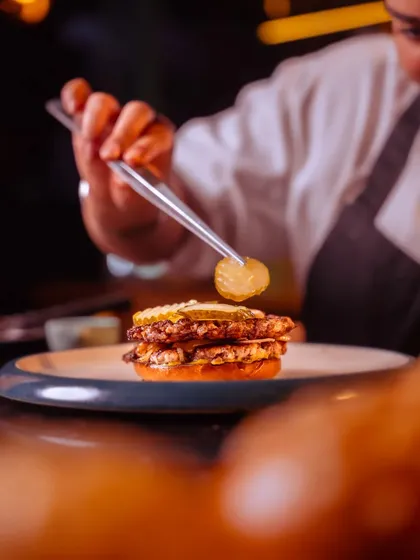 A chef carefully places a pickle on our Oklahoma-style smashed beef burger, the final touch on a culinary dream.