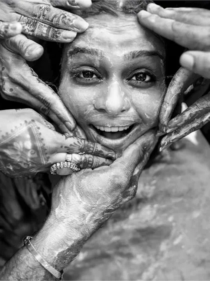 A powerful, emotional moment from a Haldi ceremony, captured in high-contrast black and white. The focus on the bride's face surrounded by hands creates a deeply impactful image.