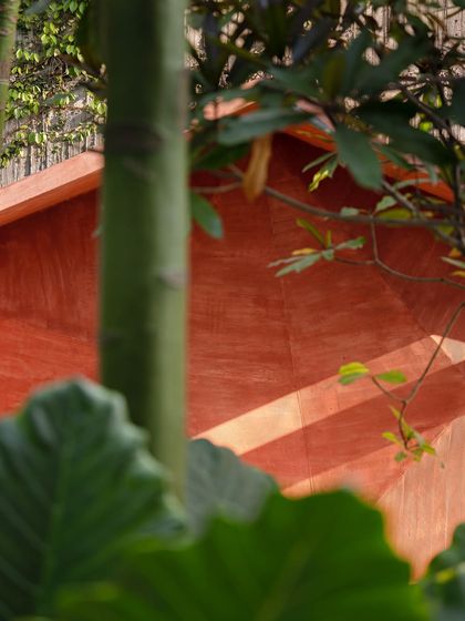 A detail shot of the red oxide sculptural bench, showing the play of light and shadow on its faceted surfaces. The texture of the finish contrasts with the soft leaves of the surrounding plants.