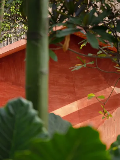 A detail shot of the red oxide sculptural bench, showing the play of light and shadow on its faceted surfaces. The texture of the finish contrasts with the soft leaves of the surrounding plants.