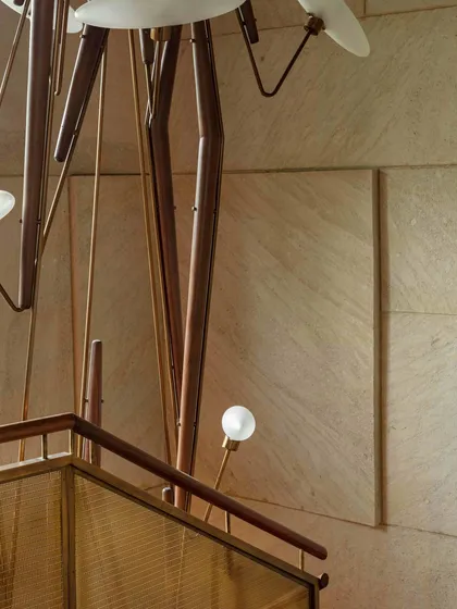 A close-up of the Klove Grasshopper light installation against a textured stone wall, highlighting the intricate details of the staircase design. The interplay of light, metal, and stone creates a dynamic visual effect.