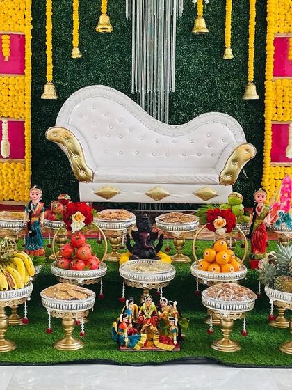 A full view of a colorful baby shower setup with a pink, yellow, and green checkered backdrop. The decor includes a white sofa and a complete arrangement of plates and dolls on an artificial grass carpet.