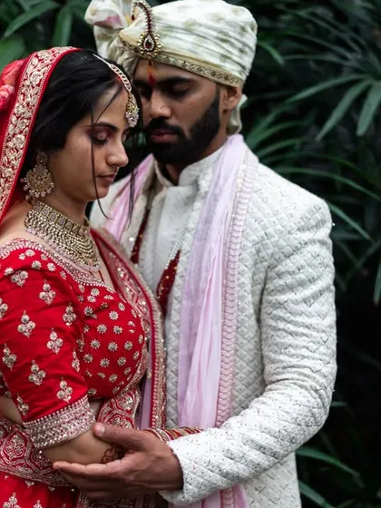 An intimate wedding portrait against a dark, natural background. The focus is entirely on the couple, highlighting their traditional red lehenga and white sherwani.