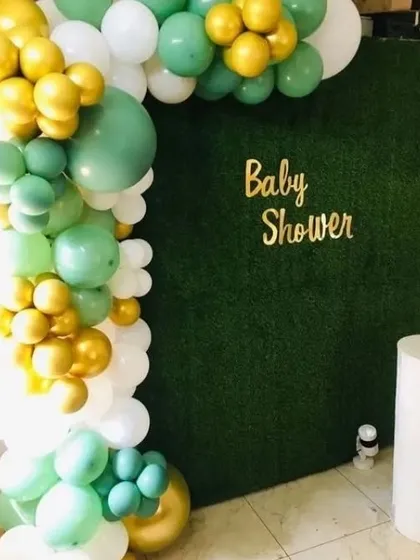 A beautiful baby shower backdrop using a faux grass wall. The wall is decorated with a 'Baby Shower' sign and a balloon garland in sage green, white, and gold.
