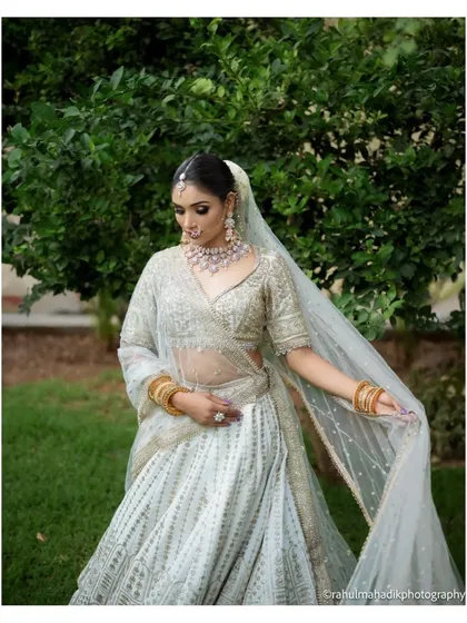 A full-length portrait of a bride in a stunning white lehenga, set against a lush green garden. Her elegant pose and the classic beauty of her attire are the focus of this timeless shot.