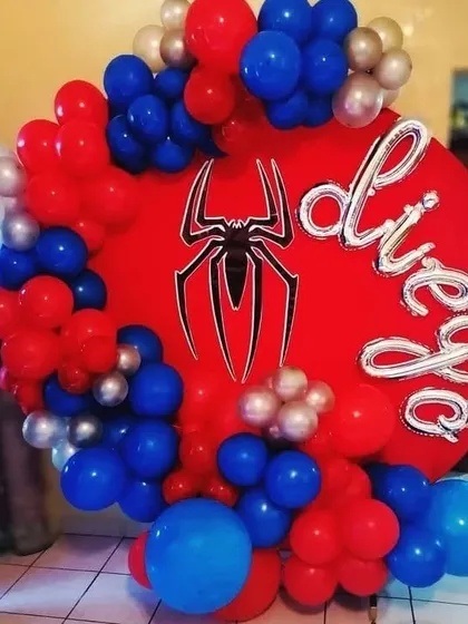 A circular backdrop with a Spider-Man theme. The red panel features the spider logo, surrounded by a garland of red, blue, and silver balloons, with the birthday boy's name in foil letters.