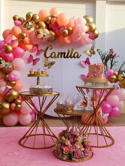 An elegant outdoor butterfly party setup with a pink and gold color scheme. The decor features a circular backdrop adorned with gold and pink butterflies, stylish dessert tables, and a beautiful balloon arch.