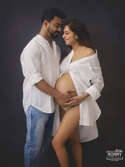 An intimate and loving studio portrait. The simple white shirt and jeans create a relaxed, natural feel, focusing on the couple's tender connection as they hold the baby bump.