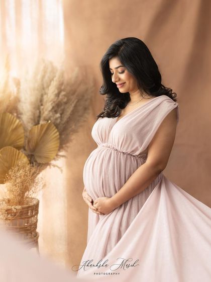 Soft light streams from the side, illuminating the mother-to-be in a flowing mauve gown against a backdrop of dried pampas grass.