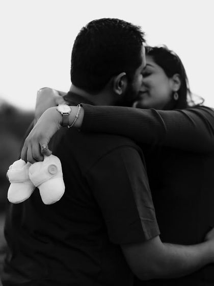 A romantic and intimate black and white photo. The couple embraces while she holds a tiny pair of baby shoes, a subtle and sweet symbol of their growing family.
