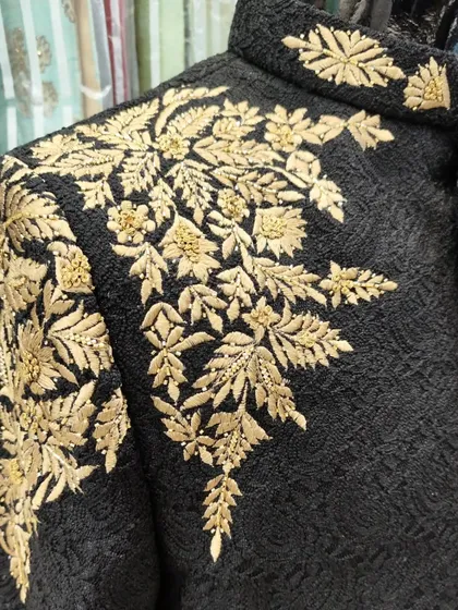 A close-up of the gold hand embroidery on a black Jodhpuri suit. The leaf-patterned threadwork is meticulously crafted to create a rich and luxurious texture.