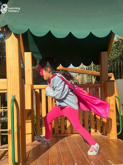 A superhero in a pink cape takes flight on the playground. Pretend play outdoors allows children to be expansive in their movements and ideas, building confidence and gross motor skills.