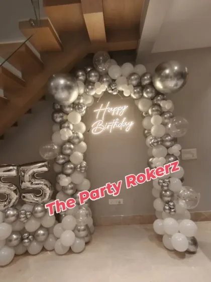 A chic balloon arch in silver and white for a 55th birthday celebration at home. The setup is placed neatly under a staircase and includes large foil number balloons.