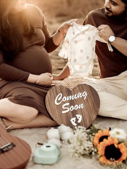 A close-up of the couple at their picnic, looking at a baby onesie together. The ukulele and "Coming Soon" sign complete this lifestyle announcement scene.