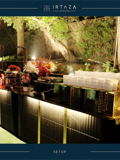 A beautiful outdoor catering setup with a natural backdrop. The warm lighting on the food counters creates an inviting glow, perfect for evening events and receptions.
