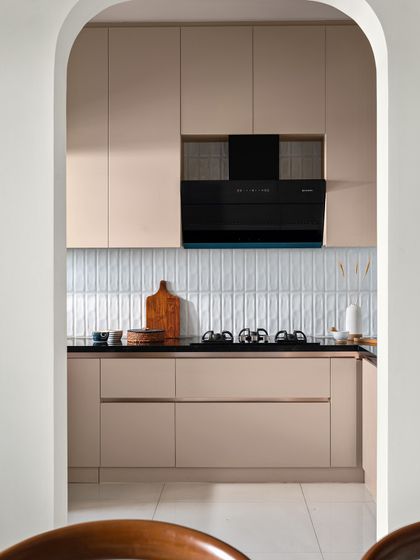 A view into a modern pastel kitchen through an elegant arched entryway. The design features beige handleless cabinets and a backsplash of vertically laid subway tiles, creating a clean and sophisticated look.