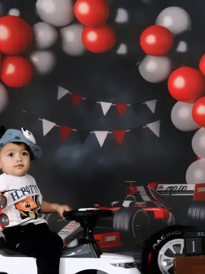 A toddler enjoys a race car themed birthday shoot, sitting in a white toy car against a backdrop of tires and checkered flags.
