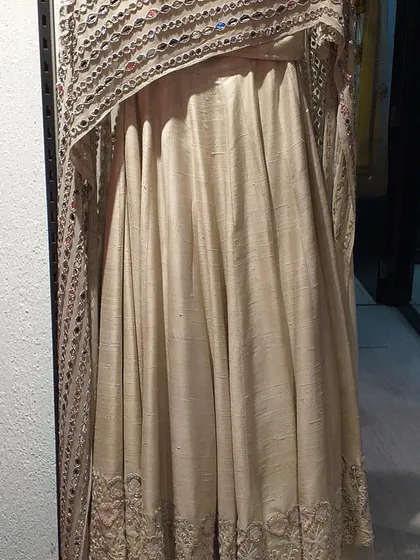 An elegant cream-colored Anarkali made from raw silk with a stunning cape-style dupatta featuring heavy mirror work. This is a luxurious and traditional piece for a major celebration.