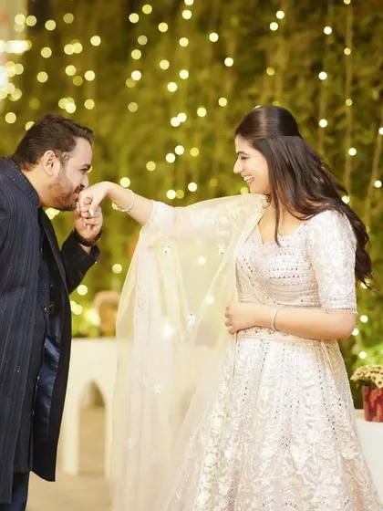 A classic romantic gesture, the groom kissing the bride's hand, captured against a backdrop of fairy lights.
