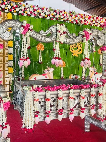 A traditional cradle ceremony setup with a silver cradle adorned with pink and white garlands against a banana leaf backdrop.