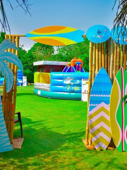 An entrance to the activity zone at the beach party, framed by bamboo poles and surfboard props. In the background, you can see a large inflatable water slide, perfect for outdoor fun.
