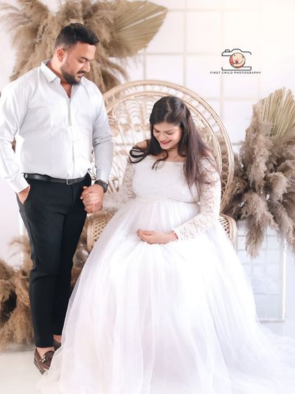 A beautiful couple's portrait from a bohemian-themed studio session. The partner holds his wife's hand as she sits gracefully in a white gown, surrounded by pampas grass.
