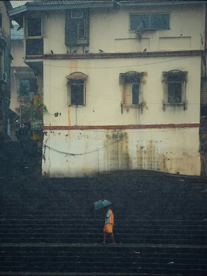 A person in saffron robes walks with an umbrella along the dark, wet steps of Banganga. The image has a grainy, film-like quality that enhances its timeless and spiritual feel.