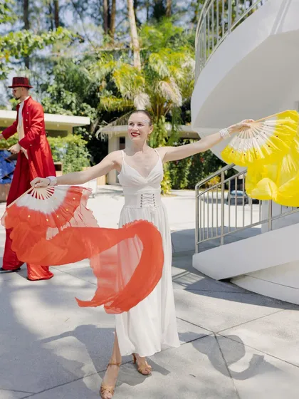 A fan dancer in a white dress with red and yellow fans, part of a coordinated troupe that brings energy and artistry to the event.