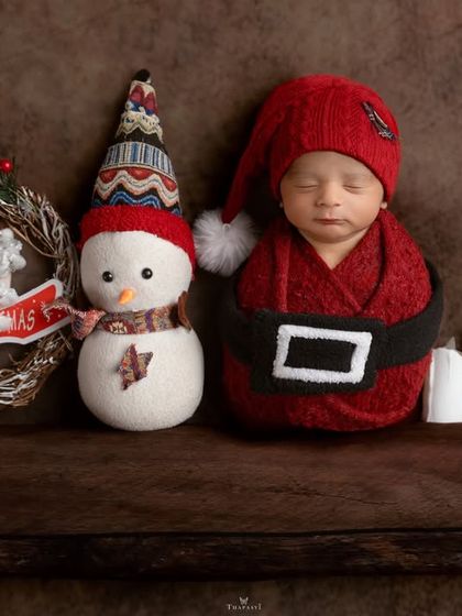 Santa's tiniest helper. A newborn dressed in a little Santa suit, sleeping on a wooden shelf next to a snowman and candles.